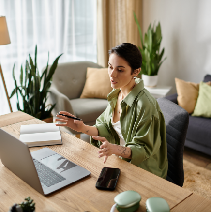A person in an olive shirt sits at a wooden desk with a laptop, gesturing while speaking during a video call.