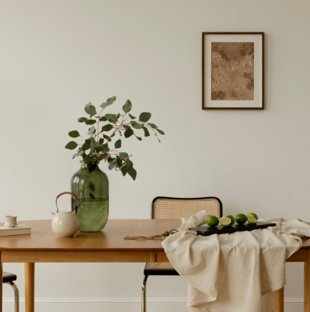 Minimalist dining room with a wooden table, green glass vase, small teapot, and a bowl of limes on a beige cloth.