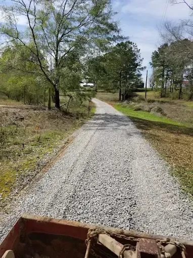 Gravel driveway curving through a grassy rural area with trees and utility poles under a cloudy sky
