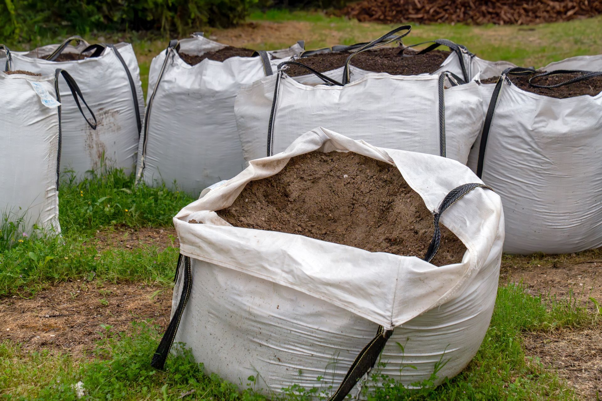 White yard waste bags filled with soil and leaves on grass, one bag open in front.