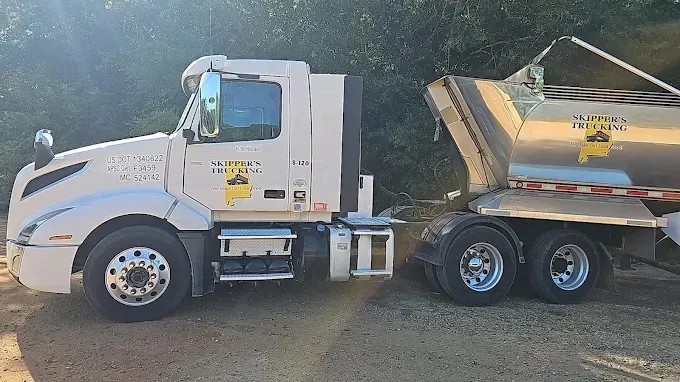 White dump truck with raised yellow trailer parked on a dirt road in a wooded area.