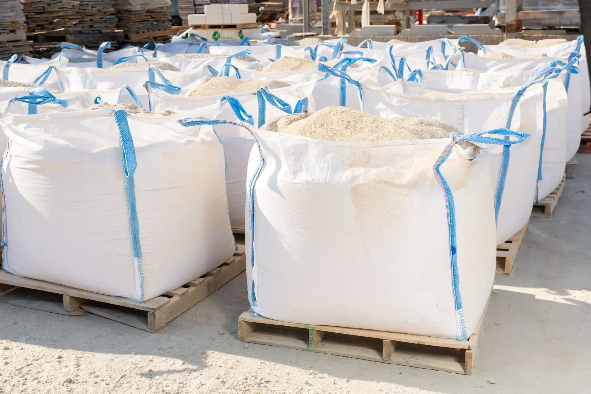 Large white bulk bags on wooden pallets outdoors, each tied with blue straps, at a storage yard