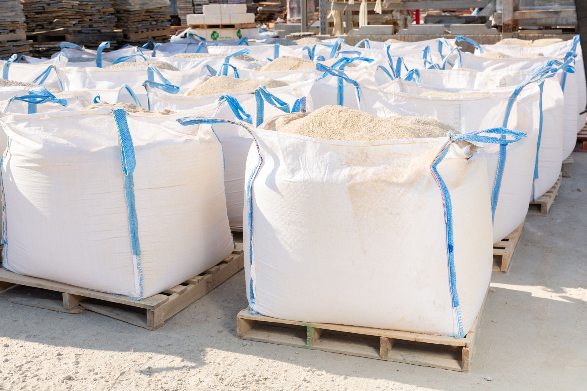 Large white bulk bags on wooden pallets outdoors, each tied with blue straps, at a storage yard
