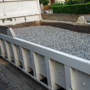 Gray gravel pile in a white dump trailer beside a building wall and driveway.