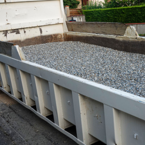 Gray gravel pile in a white dump trailer beside a building wall and driveway.