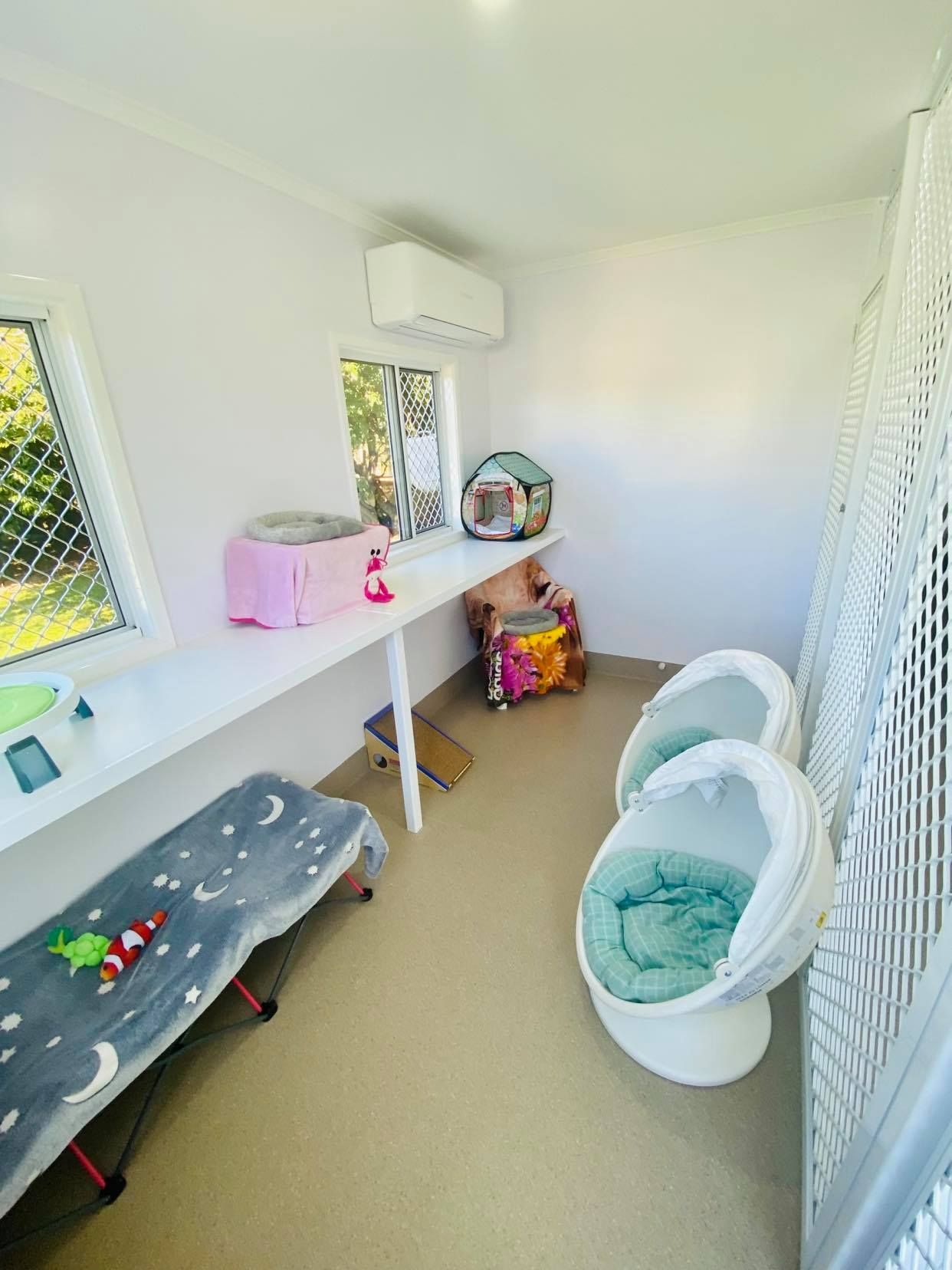 Cat boarding room: white walls, beds, toys, windows. Beige floor, air conditioner — Whispurrs Cat Retreat in Beerburrum, QLD