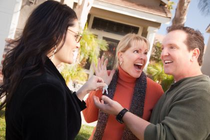 Three people smiling and talking outside a house, one holding a small object.