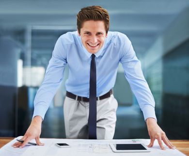 Smiling man in a blue shirt leaning on a desk in a bright office, looking at the camera.