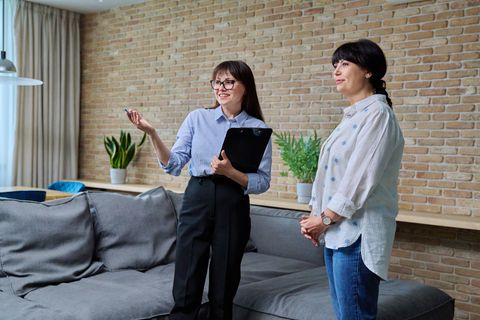Two people standing in a living room, one holding a tablet and gesturing beside a gray sofa.