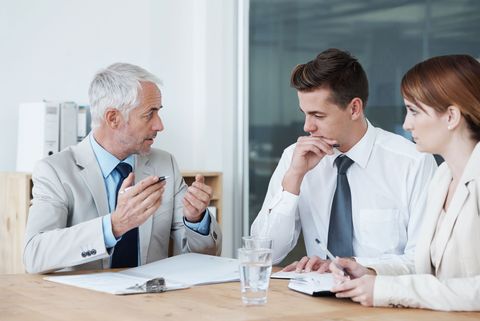 Three coworkers in a meeting, one man speaking while two others listen at a table with notes and a glass of water