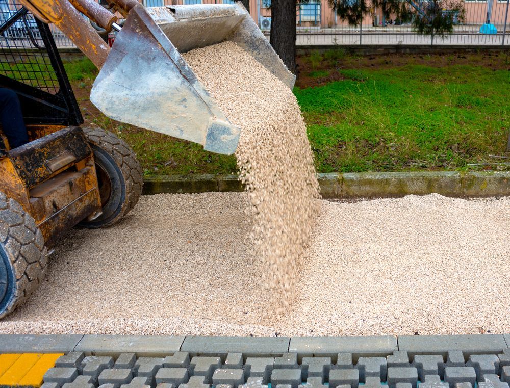 A Small Loader Pours Tan Gravel Onto a Sidewalk Next to a Grassy Area and a Tree — Boland Contracting In Wellington, NSW
