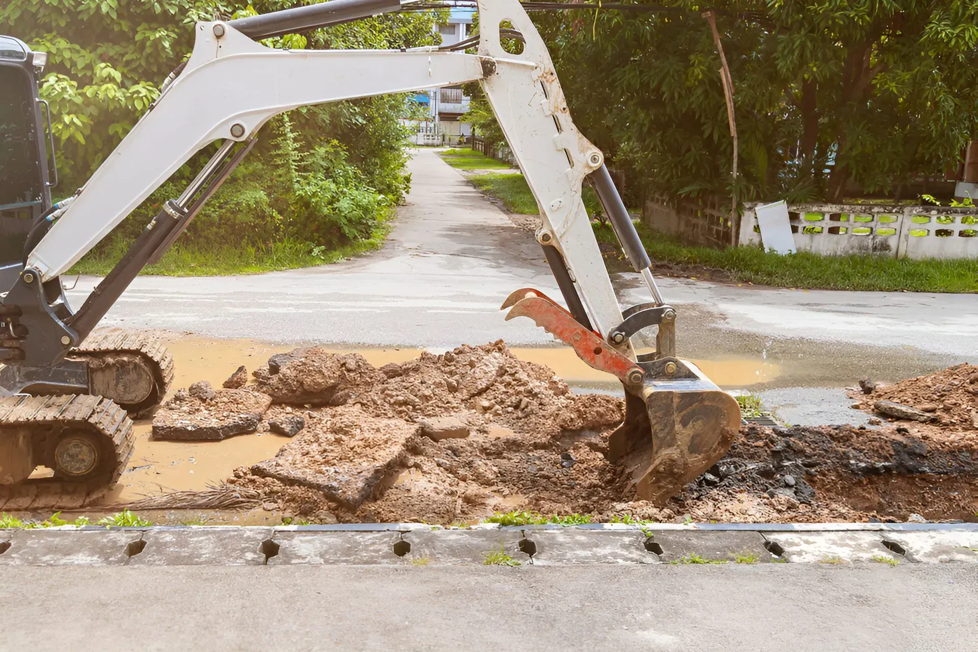 A Small White Excavator Digging Up Pavement on a Residential Street — Boland Contracting In Parkes, NSW