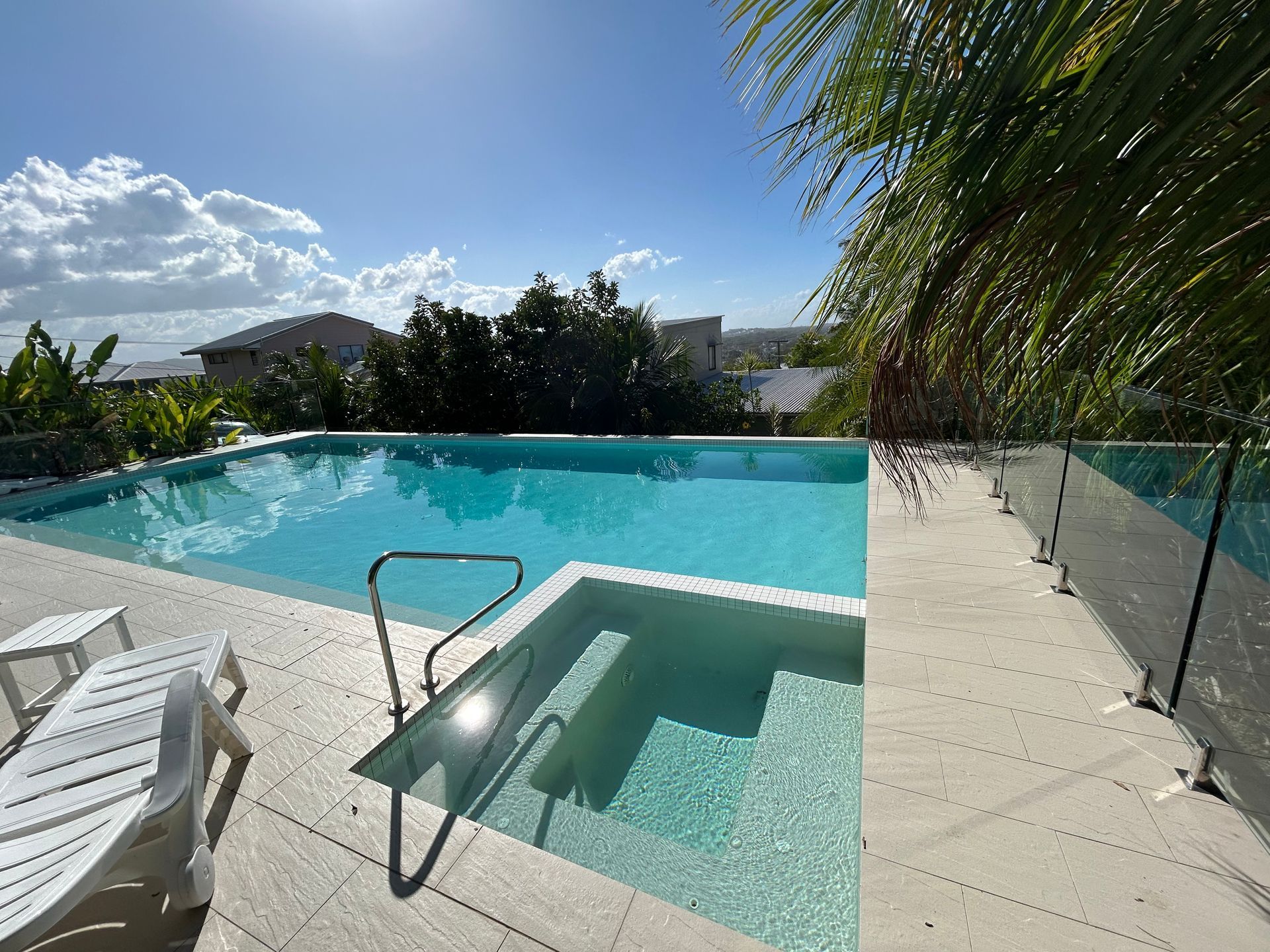 Sunny Backyard Pool With Palm Trees, Lounge Chair, and Shallow SPA Area Beside a Glass Fence — Bromage Pool Heating in Palmview, QLD