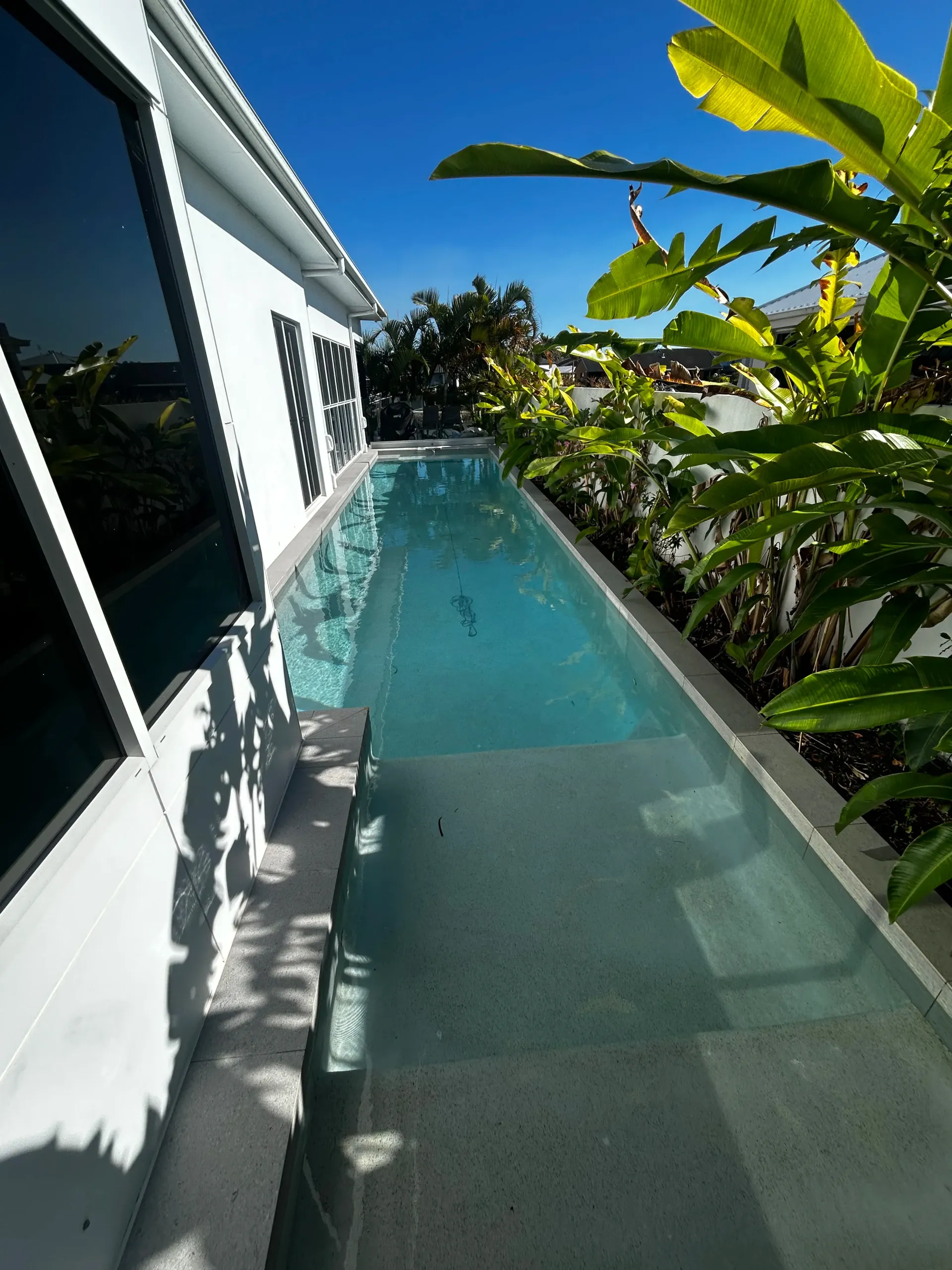Narrow Turquoise Pool Beside a White House, With Tropical Plants and Blue Sky Overhead — Bromage Pool Heating in Palmview, QLD
