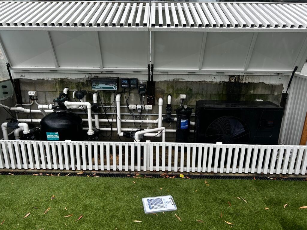 Outdoor Utility Equipment Behind a White Fence, With Pipes and Machines Under a Corrugated Metal Roof — Bromage Pool Heating in Caloundra, QLD
