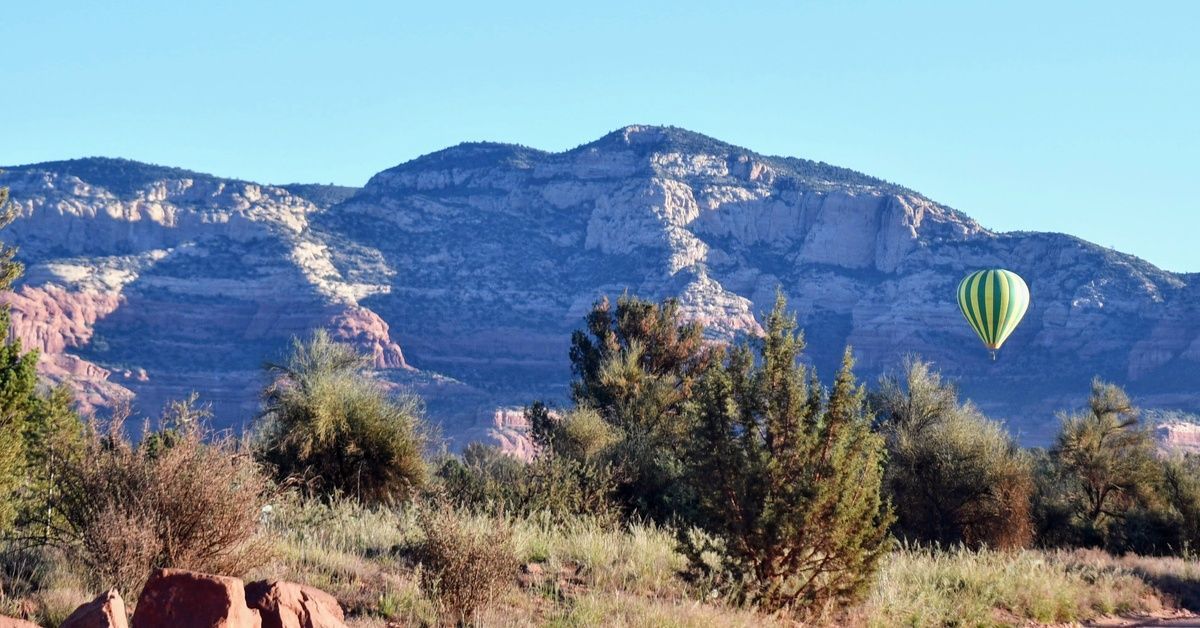 Three hot air balloons fly over a Sedona, AZ landscape. Two balloons are green and yellow, and the other looks rainbow-colored.