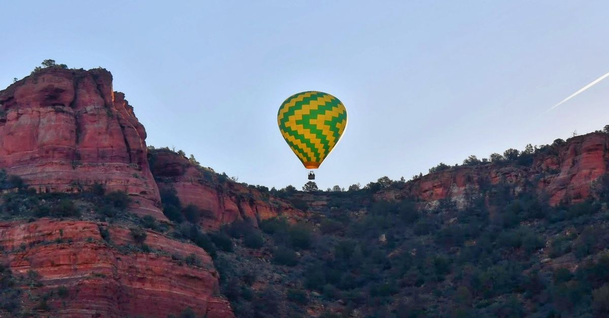 A hot air balloon in the air with a clear, blue sky behind it. A towering rock formation covers the area below the balloon.