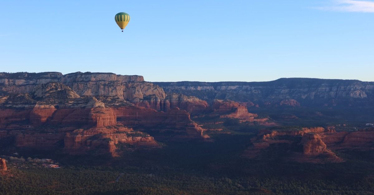 Hot air balloon floats above Sedona’s shadowed red rocks. Blue sky stretches over the canyon