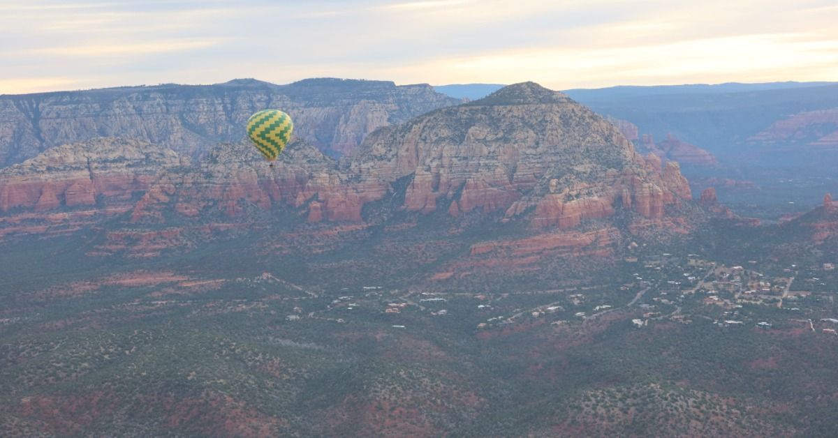 A yellow-green balloon glides before Sedona’s pale cliffs. Soft sunrise haze spreads across the red rock valley.