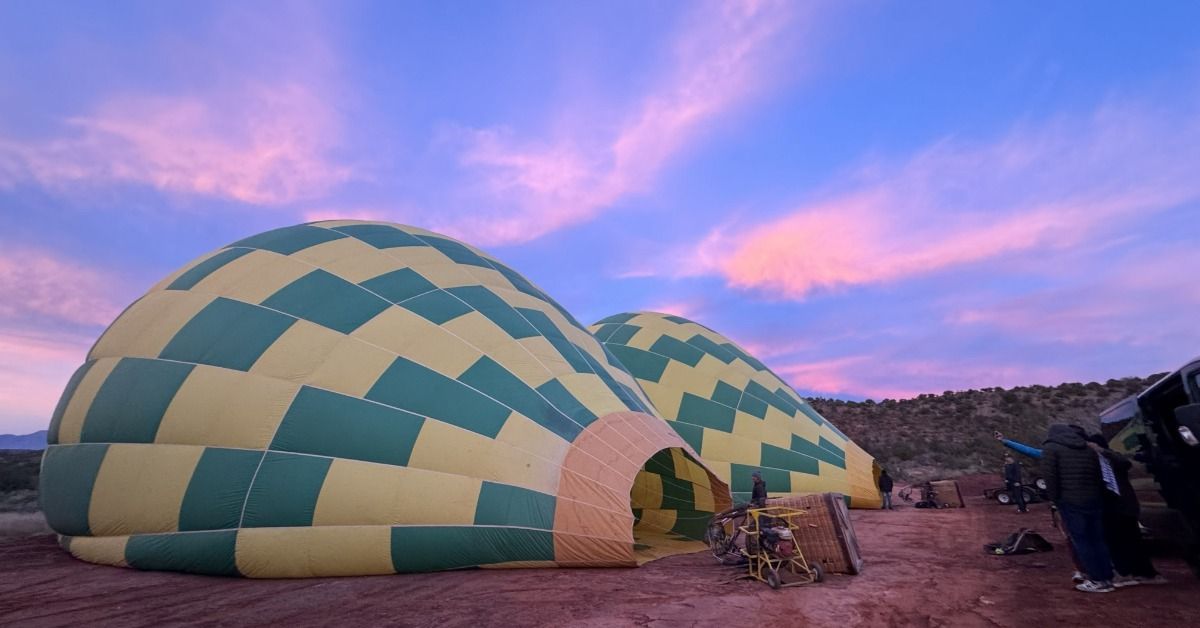 Two striped balloons inflate on red desert ground at dawn. Pink sunrise clouds glow above the launch area.