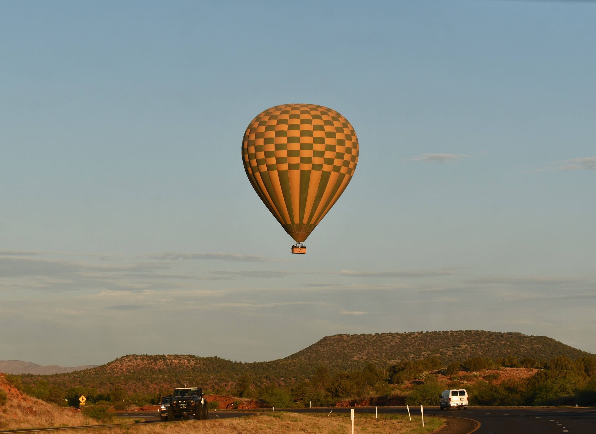 A yellow-and-green hot air balloon floats above a desert highway at dawn. Low red hills stretch bene