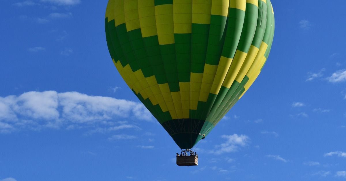 A green-and-yellow hot air balloon rises above a rocky desert ridge. Small white clouds scatter across the deep blue sky.