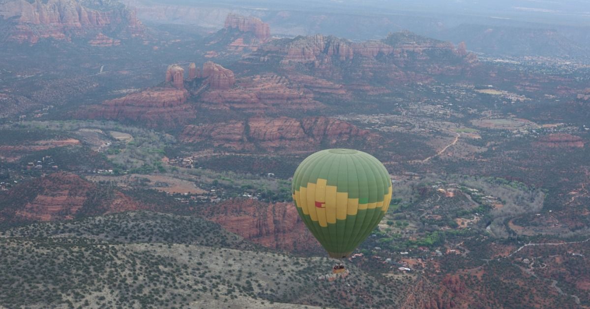 A green hot air balloon drifts over Sedona’s red rock canyons. Roads, homes, and layered mesas spread across the valley.