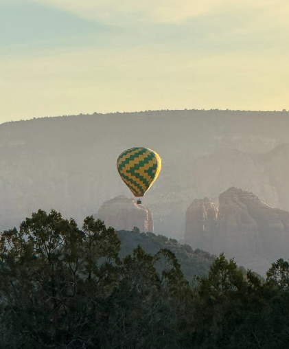 A hot air balloon with a green and yellow checkered pattern floats above the desert trees and peaks.