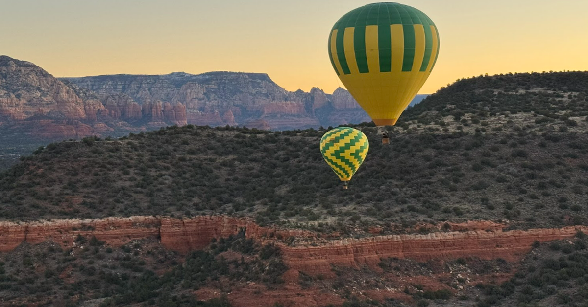 Two yellow and green hot air balloons are floating above a terrain of red rocks and desert plants.