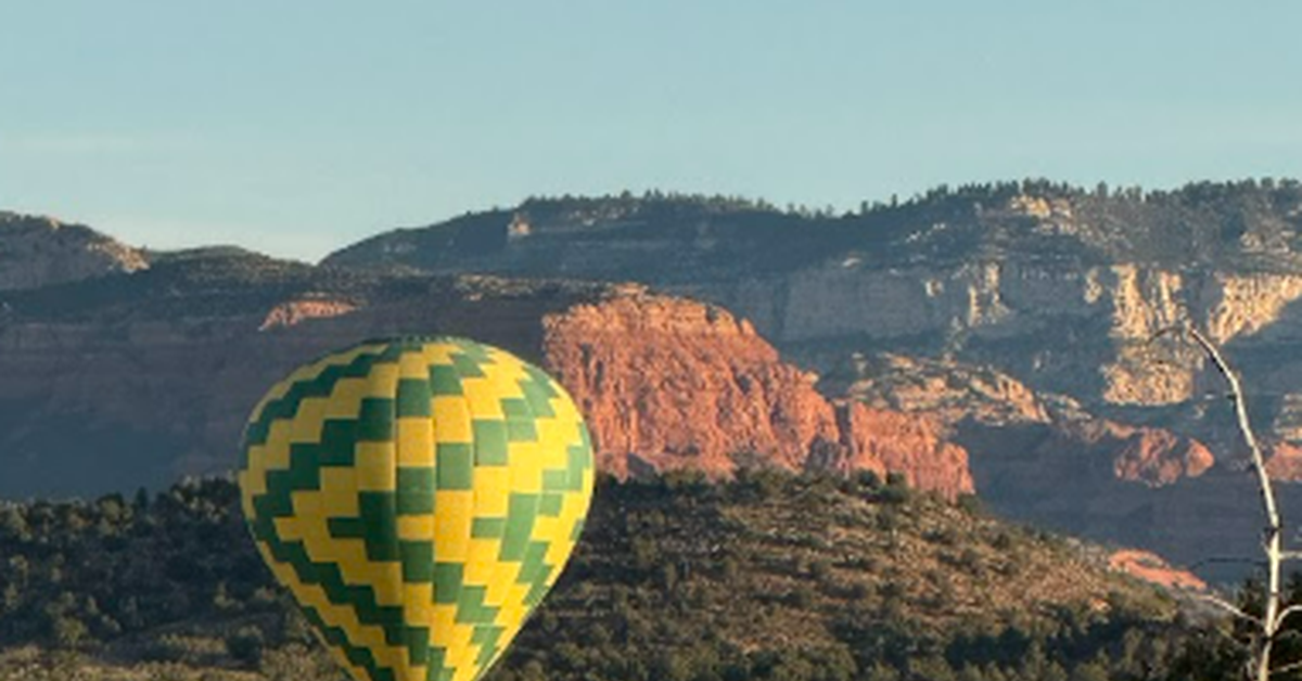 A yellow and green hot air balloon is low to the ground in a desert region. It's surrounded by red rocks and plants.