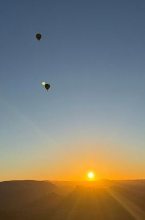 Two hot air balloons are floating above the red rocks of Sedona. The sun and the moon are both in the sky behind them.