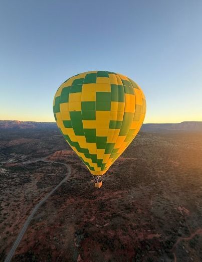 A green and yellow hot air balloon is floating in the sky above a desert region. The sun illuminates the landscape.