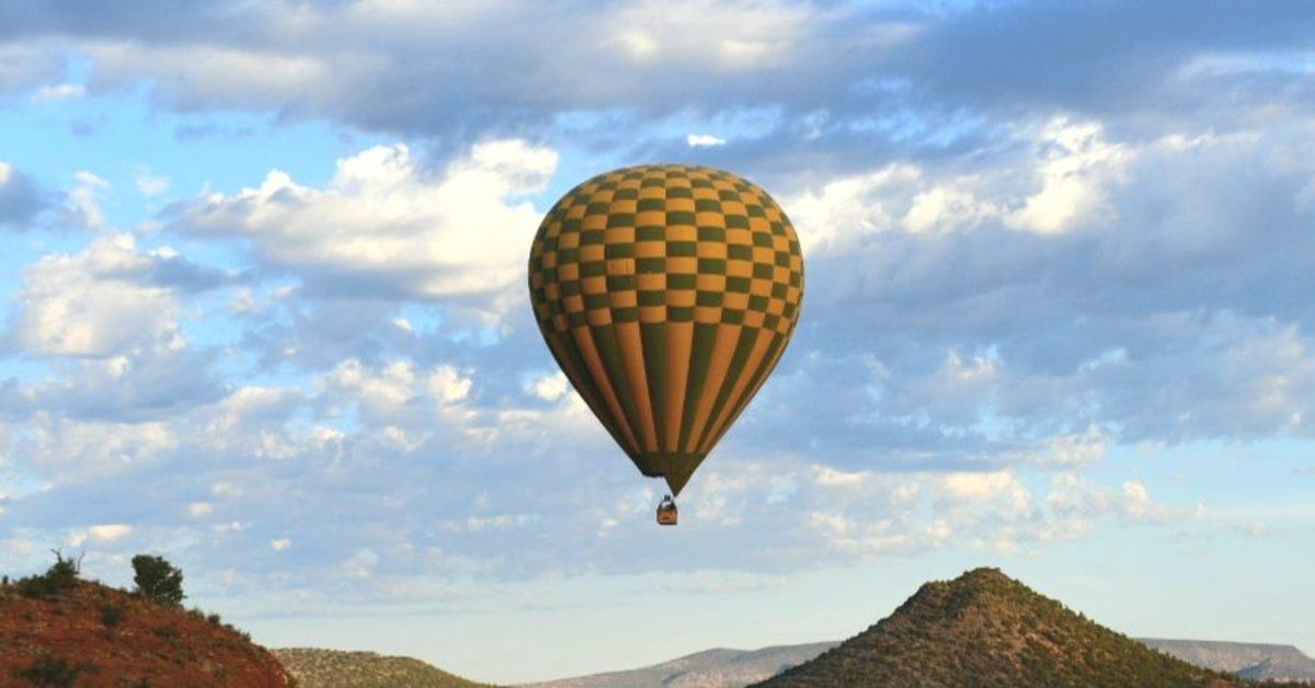 A checkered hot air balloon floats above red rock hills and scenic desert views beneath clouds.