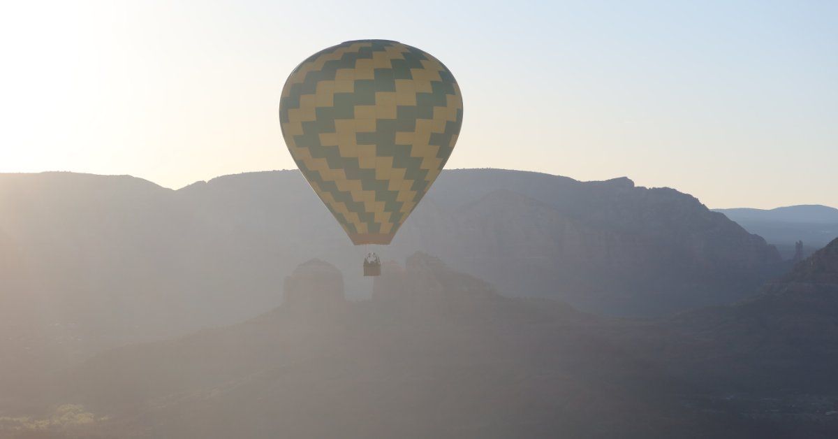 A patterned hot air balloon rises over misty canyon landscapes at sunrise, silhouetted against soft morning light.