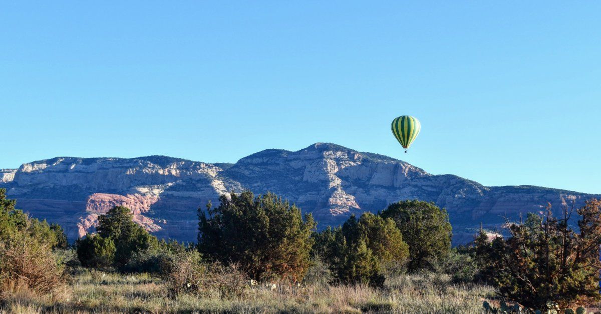 A hot air balloon drifts above red rock formations and desert brush under a clear blue sky.