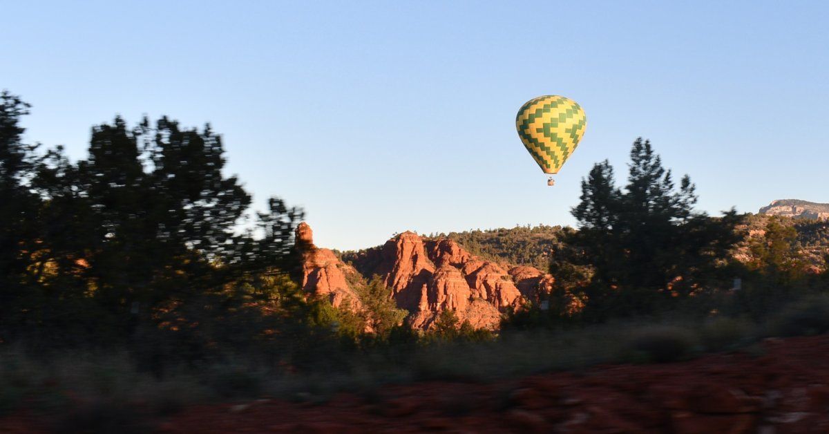 A hot air balloon drifting above red rock formations and pine trees, showcasing a scenic desert landscape at dawn.