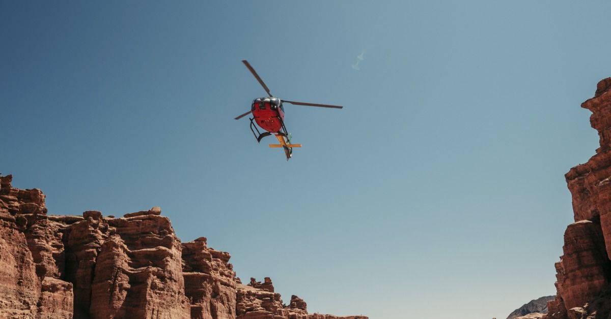 A red sightseeing helicopter flying above a rugged desert canyon cliffs beneath a clear blue sky.