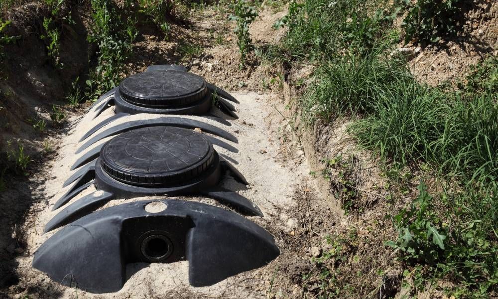 A black septic tank with two covers partially imbedded in the ground with sand on top and the drain 