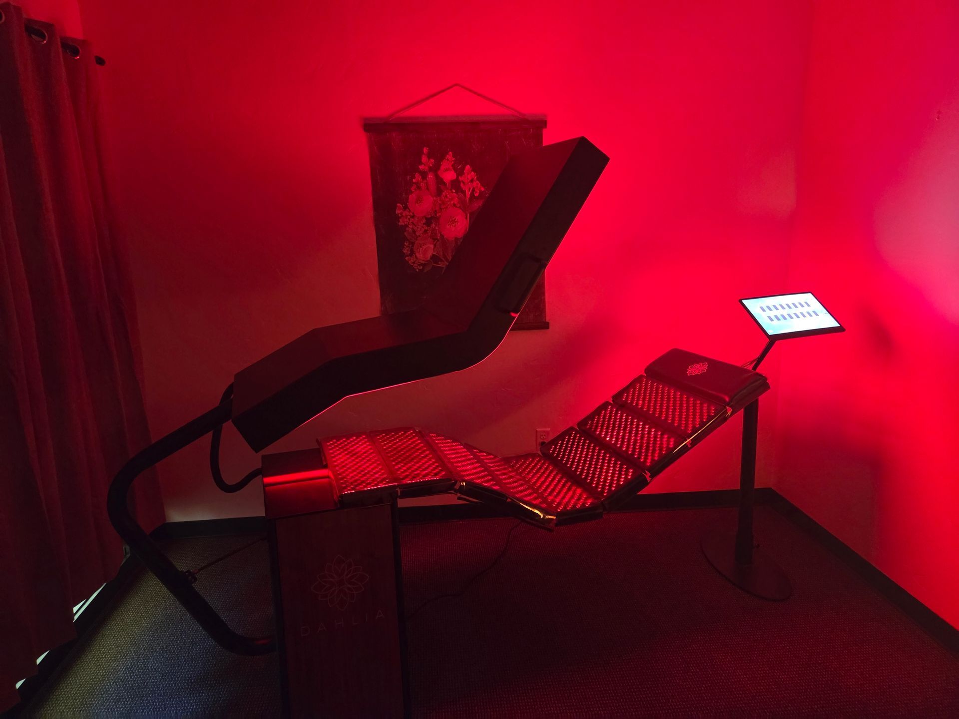 A red-lit room contains a red light therapy bed with a raised overhead panel and a small control screen on a stand.