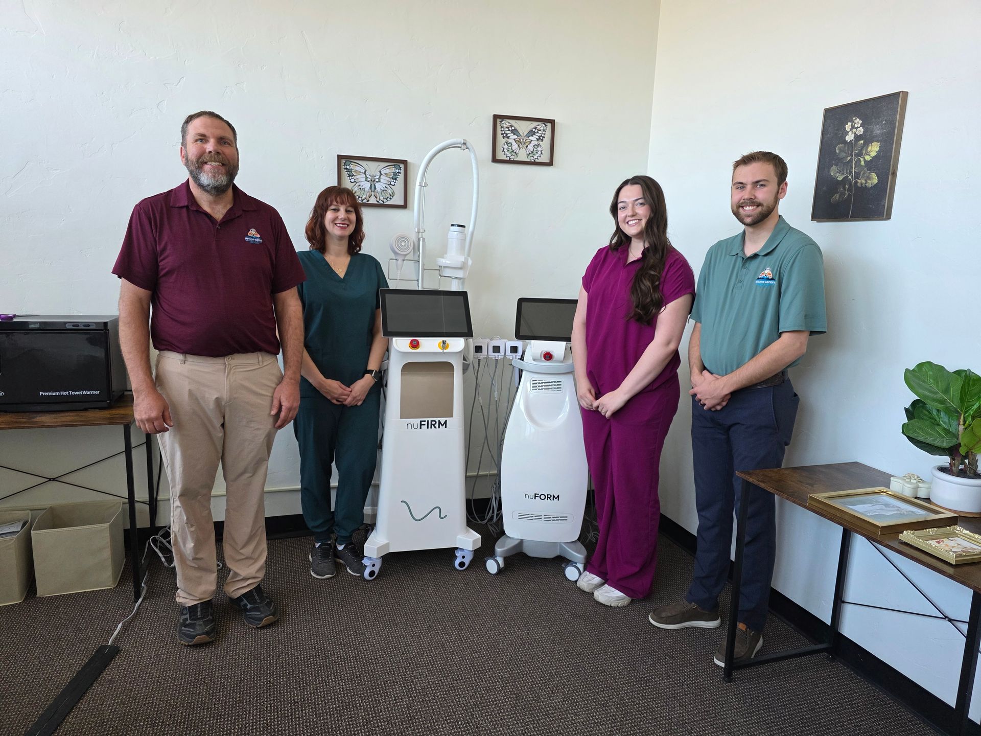 Four people stand smiling in an office with two medical laser machines, framed art on the wall, and wooden furniture.