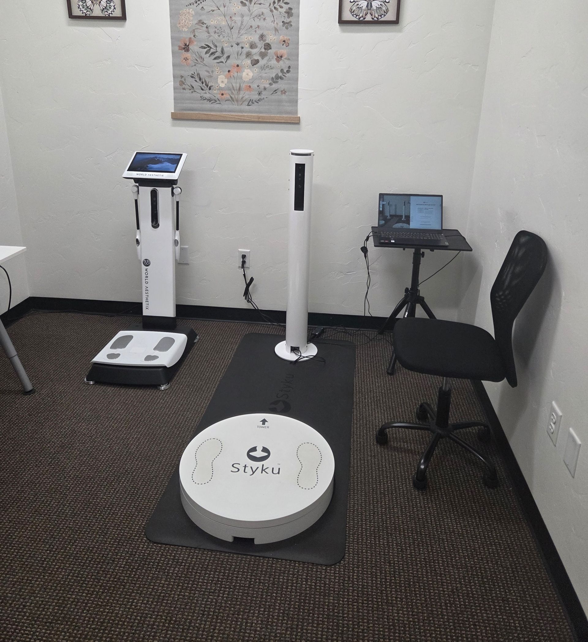 Medical room with two body composition scales, a tripod-mounted laptop, and a black office chair on a patterned carpet.