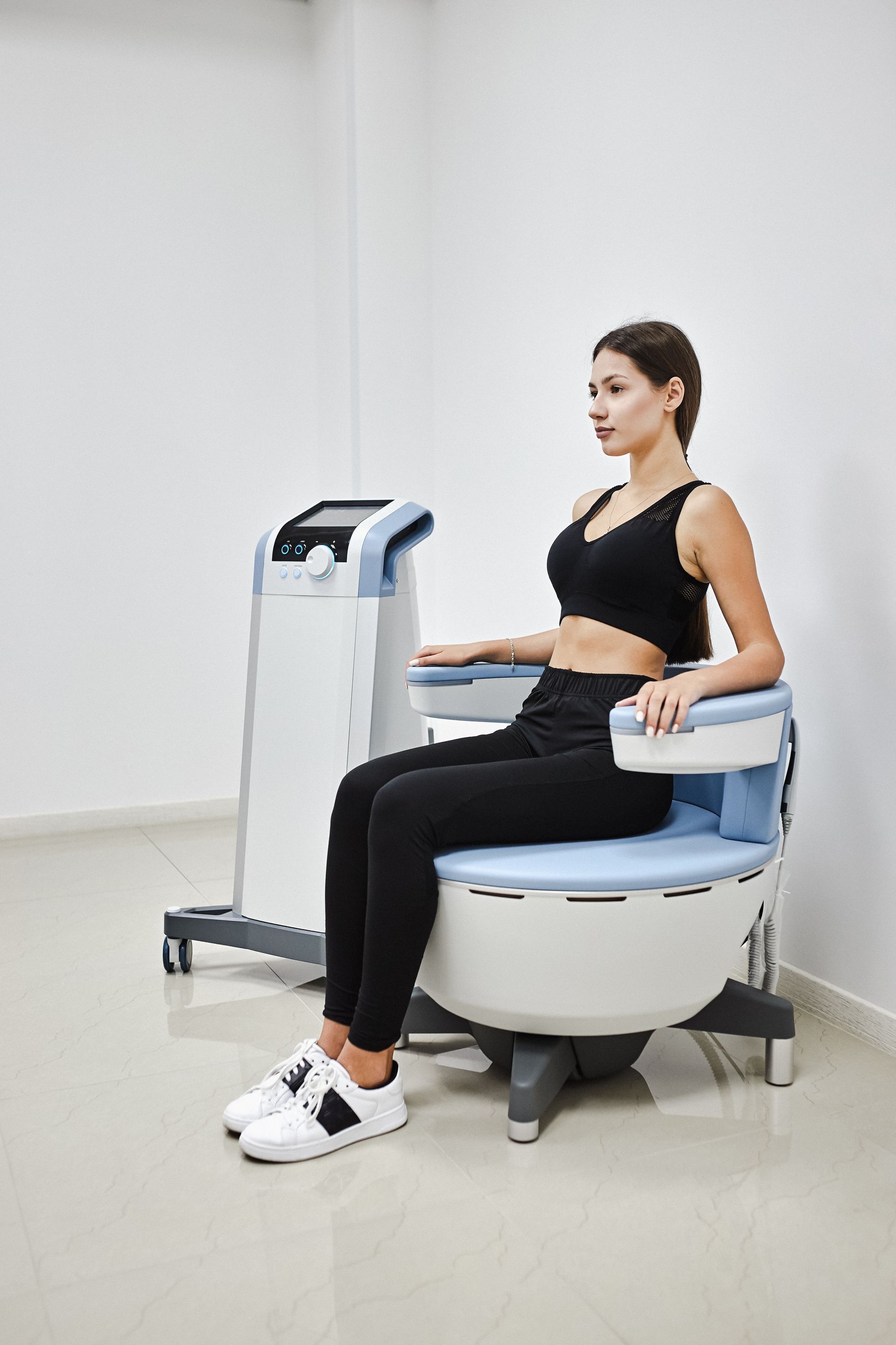 A person sits on a medical pelvic floor exercise chair next to a control console in a bright, clinical room.