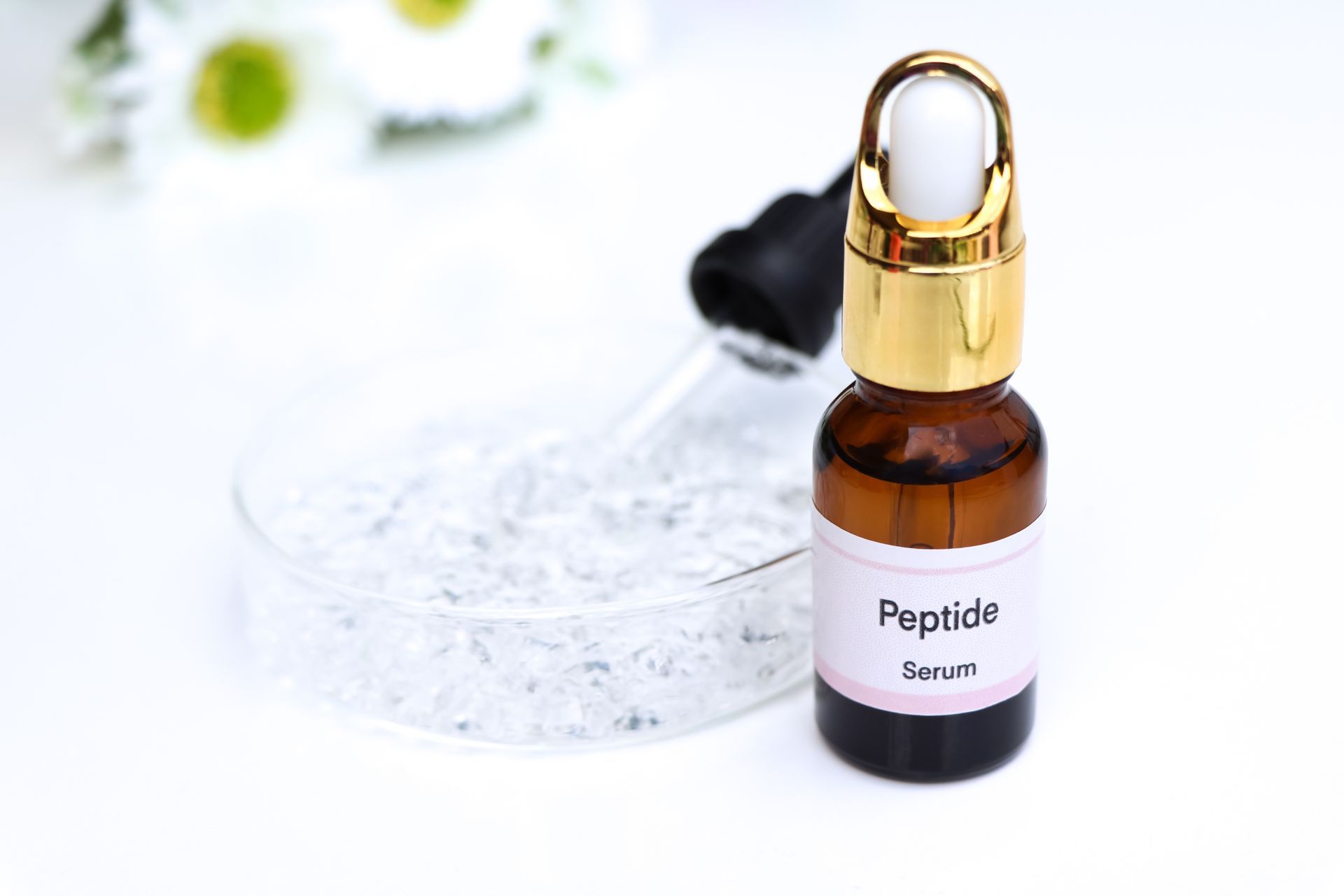 A bottle of peptide serum with a gold dropper sits next to a glass dish of clear beads on a white surface.