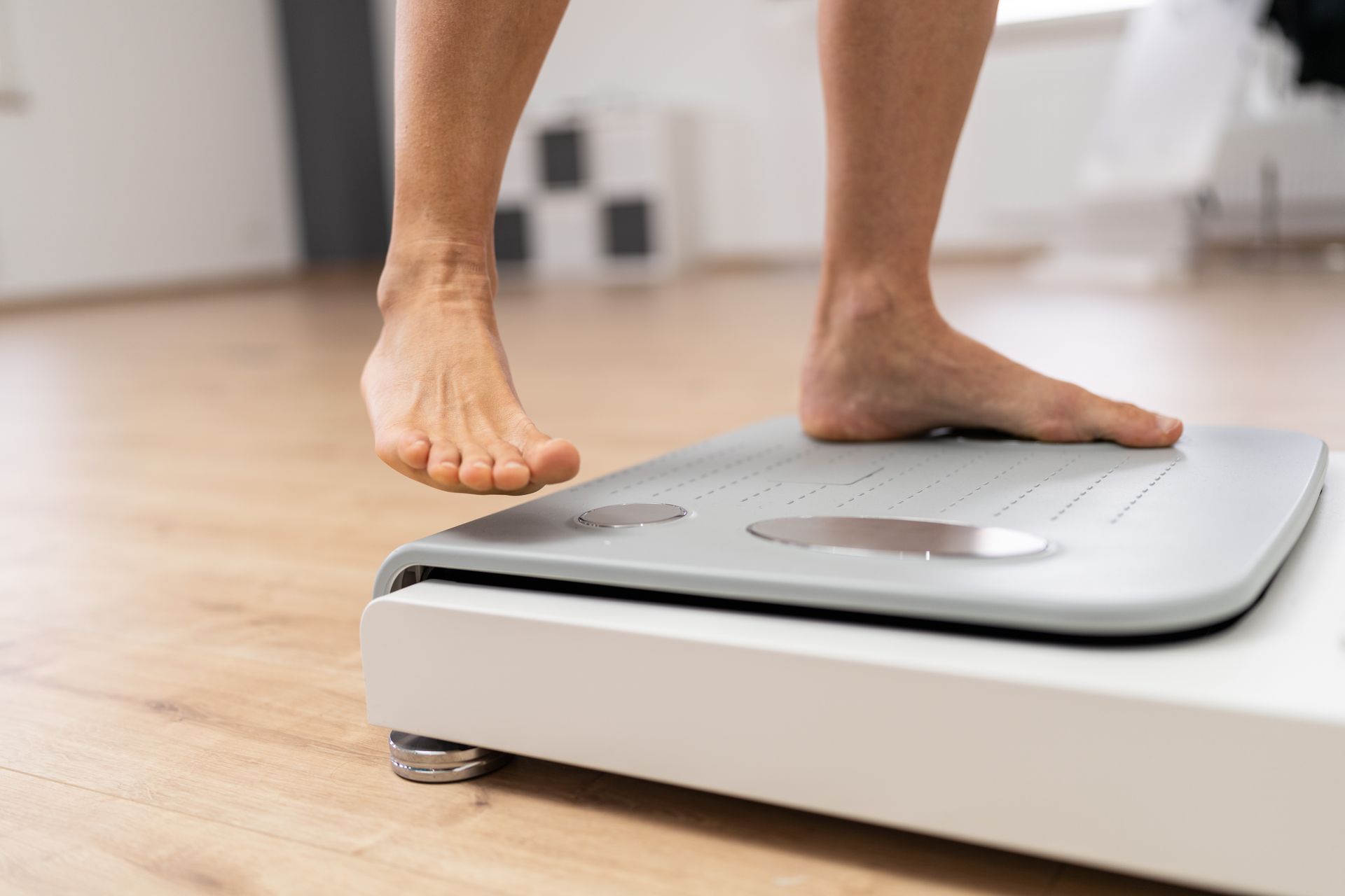 A person balancing on one foot while using a white digital scale on a wooden floor.