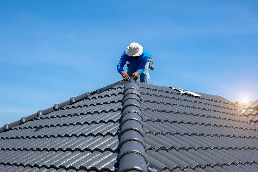 Roofer in blue shirt and white hat working on gray tile roof against a blue sky — Mullum Property Services in Ballina, NSW