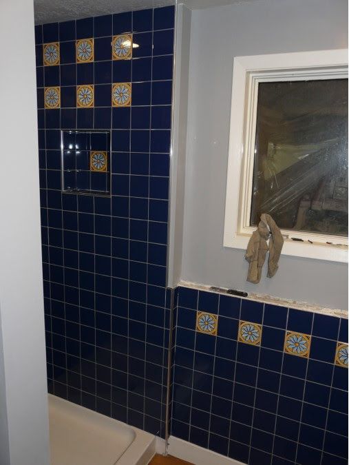 A tiled bathroom wall features deep blue square tiles with decorative yellow and white floral accents and a recessed niche.