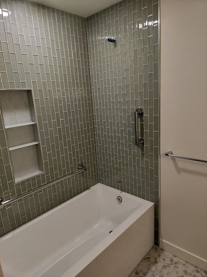 Bathtub shower area featuring green vertical mosaic tile walls, a built-in shelf, grab bars, and a white tub.