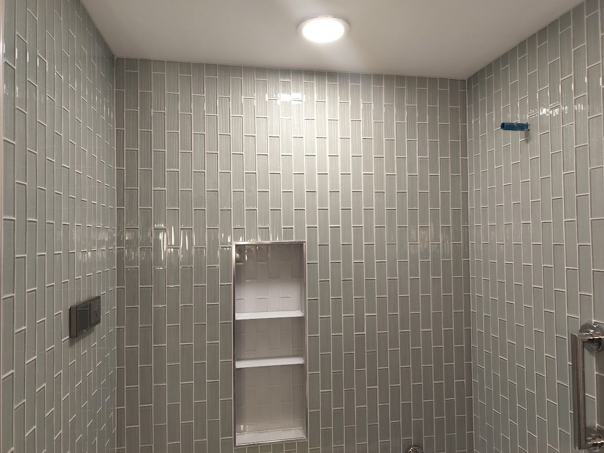 A tiled shower with light gray, vertically oriented rectangular tiles, a recessed shelf, and a ceiling light.