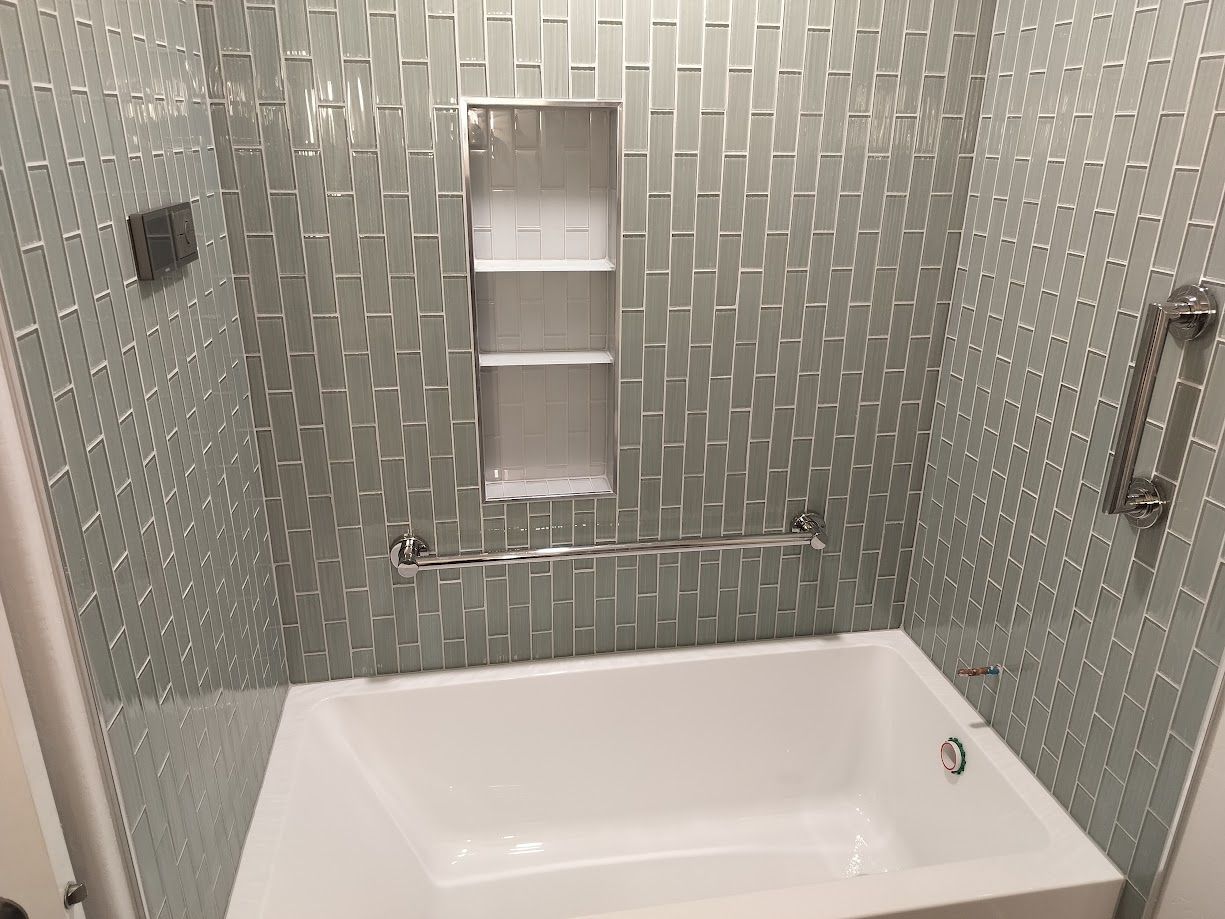 A white bathtub with silver grab bars installed on a light-grey tile wall featuring a built-in recessed shelf.