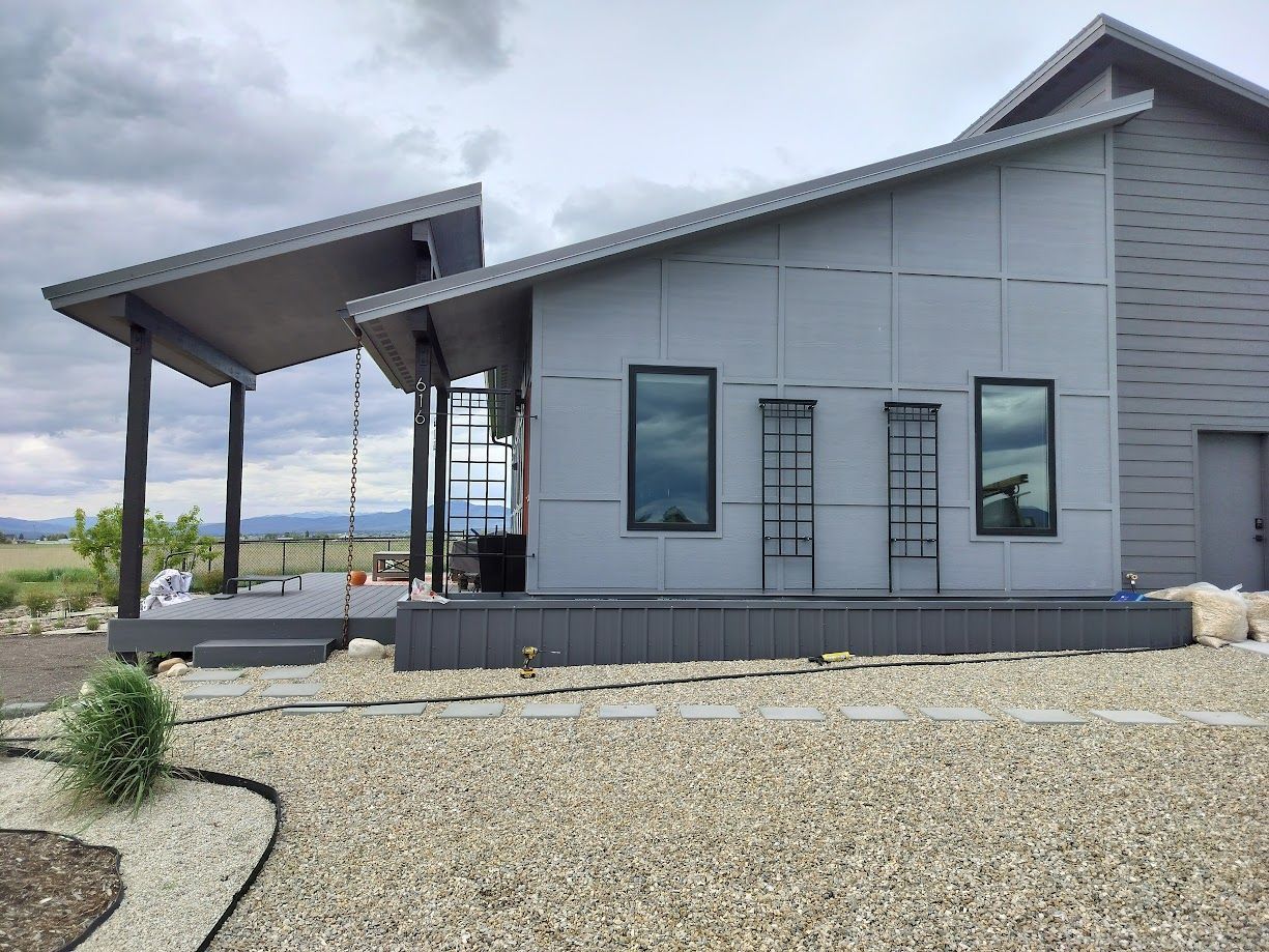 Modern gray metal house with a sloped roof, covered deck, large windows, and a gravel yard under a cloudy sky.